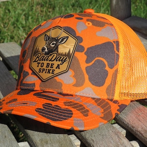 May include: An orange and brown camouflage baseball cap with a mesh back. The front features a tan hexagonal patch with a graphic of a deer head and the text "Bad Day TO BE A SPIKE" printed in black lettering across the center of the patch.