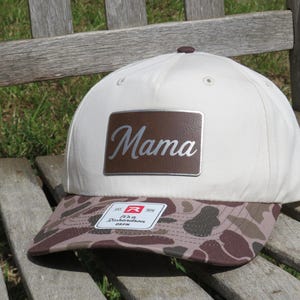 May include: A cream-colored baseball cap with a brown camouflage-patterned brim. A rectangular brown patch is centered on the front panel, featuring the word Mama in a silver script font. A small white sticker is attached to the brim.