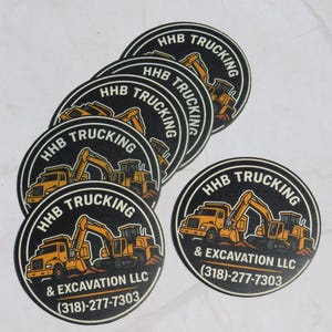 May include: A stack of circular black stickers featuring a yellow construction truck and excavator graphic. The text reads HHB TRUCKING & EXCAVATION LLC with the phone number 318-277-7303 printed in white and yellow lettering on the circular surface.