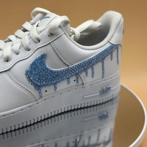 May include: White leather sneakers with a blue rhinestone swoosh and drip design.