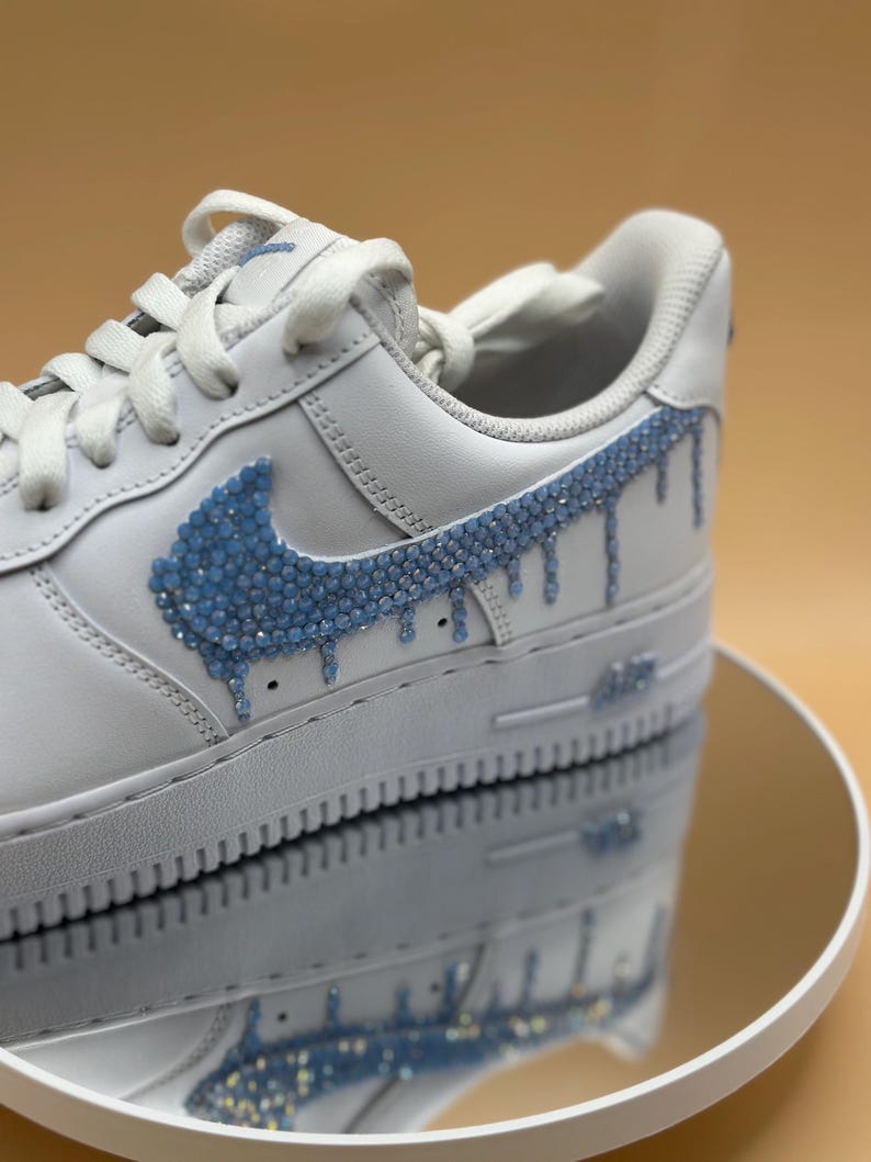 May include: White leather sneakers with a blue rhinestone swoosh and drip design.