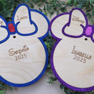 May include: Two wooden Mickey Mouse-shaped ornaments. One has a blue glitter outline and a red glitter bow, with the name "Sophia" and the year "2025" engraved. The other has a purple glitter outline and bow, with the name "Isabella" and the year "2025".