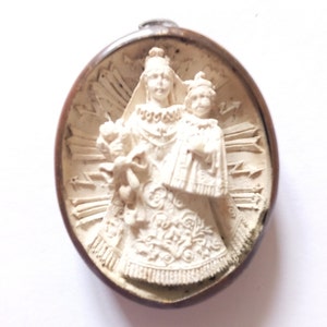 Puede incluir: Un colgante de cerámica blanca con una escultura en relieve de la Virgen María y el niño Jesús. El colgante es circular y tiene un marco de metal marrón.
