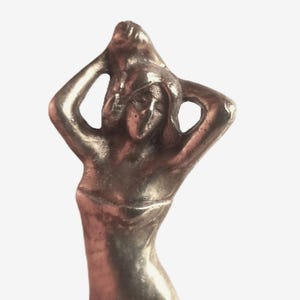 May include: A silver-coloured sculpture of a person with arms raised above the head. The figure has a smooth, flowing form, with details suggesting hair and facial features. The sculpture is set against a plain white background.