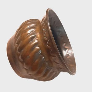 Peut inclure: Un pot en métal de couleur cuivre avec une large ouverture et un corps arrondi. L'extérieur présente une série de crêtes verticales en relief autour du centre et une bande d'indentations triangulaires répétées près du bord du récipient.