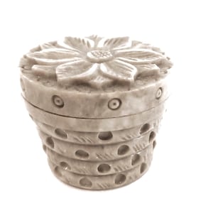 May include: A light grey, carved trinket box with a flower-shaped lid. The box has a cylindrical shape with horizontal bands and circular cutouts. The lid features detailed petal carvings. The box is made of a stone-like material.