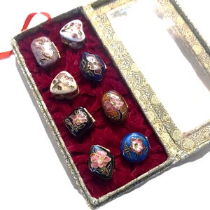 May include: A collection of seven small, hinged metal trinket boxes with colorful enamel floral designs in various shapes, including heart, oval, and round, displayed inside a rectangular box lined with deep red velvet and a clear lid.