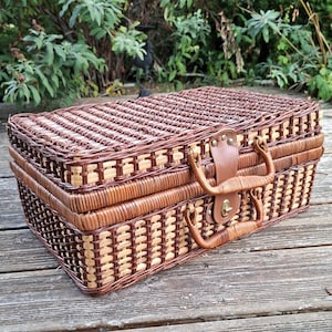 Puede incluir: Una cesta de picnic rectangular hecha de mimbre trenzado en color marrón oscuro y beige claro. La cesta cuenta con una tapa abatible, un asa trenzada resistente y un cierre central de correa de cuero sintético marrón con un pestillo giratorio de metal dorado.