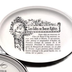Puede incluir: Una bandeja de servicio ovalada de cerámica blanca con una ilustración negra de una cabeza de vaca en un marco ornamentado y el texto Les Cotes de Boeuf Roties, seguido de una receta francesa para asar carne con referencia de 2 kg.