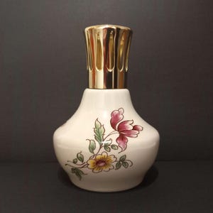 May include: Cream-colored ceramic fragrance lamp with a gold-colored metal top. The lamp features a floral design in shades of red, yellow, and green. The lamp has a bulbous body and a decorative top.