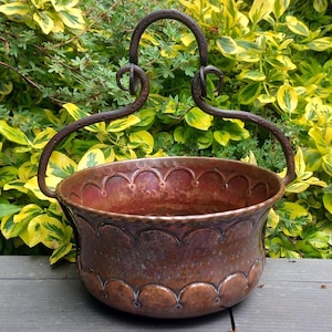 Puede incluir: Una olla redonda de cobre con un acabado metálico oscuro martillado y un borde festoneado decorativo. La olla presenta un intrincado patrón floral en relieve en el exterior e incluye un asa de hierro alta y arqueada con detalles.
