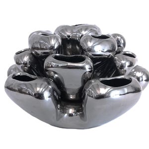 May include: A metallic silver ceramic flower frog or vase featuring multiple rounded, bulbous openings arranged in a tiered, organic cluster. The surface is highly reflective and smooth, with a glossy finish that catches light across its curves.