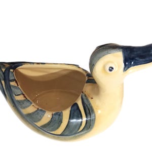Può includere: Un vaso in ceramica a forma di anatra con corpo color crema e dettagli dipinti a mano con pennellate blu sulle ali e sulla testa. L'anatra presenta un becco largo e piatto, un centro cavo per piantare e una finitura liscia e lucida.