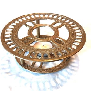 May include: A gold-colored metal candle warmer with a circular, openwork design. The warmer has a decorative pattern and a small candle is lit inside. The candle warmer is on a white surface.