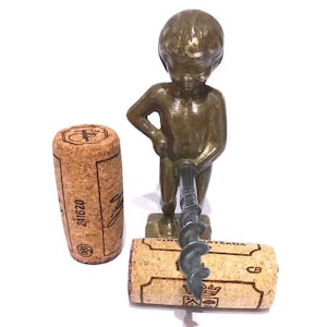 Könnte beinhalten: Ein bronzefarbener Metallkorkenzieher in Form einer stehenden Figur, positioniert mit einer nach vorne ragenden Metallspirale. Zwei hellbraune zylindrische Weinkorken sind vorhanden, einer steht aufrecht und einer liegt horizontal unter der Spirale.