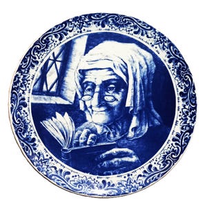 May include: A round, blue and white decorative plate with a detailed illustration of an elderly person reading a book. The plate has a detailed floral border design. The image is in a Delftware style.