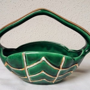 May include: A deep green ceramic basket featuring a high arched handle and a textured exterior with raised white and gold-toned lines forming a chevron pattern. The bowl has a glossy finish and a wide, open top with a curved, scalloped rim design.