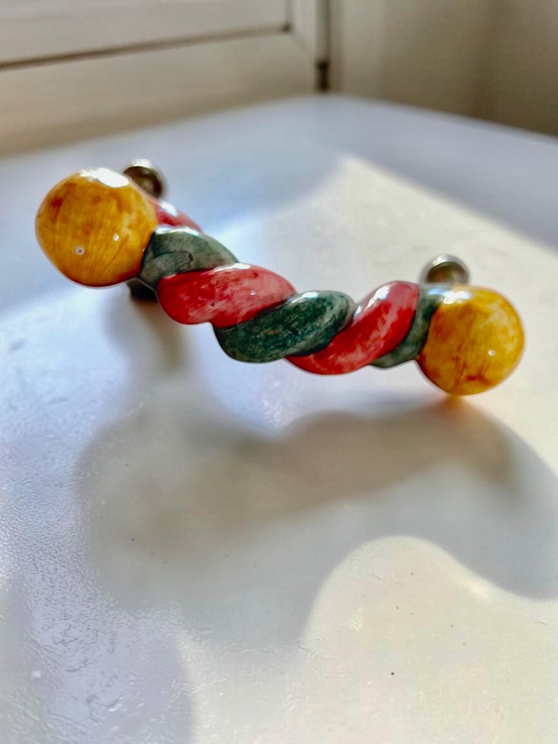 May include: A cabinet handle featuring a twisted rope design with alternating segments of yellow, red, and green. The handle has rounded ends and two silver-toned metal mounting points on the back, creating a curved shape for gripping.