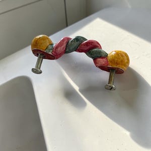 May include: A ceramic drawer pull featuring a twisted rope design with alternating segments of yellow, red, and dark green glaze. The handle has two metal mounting screws attached to the underside, extending outward for installation purposes.