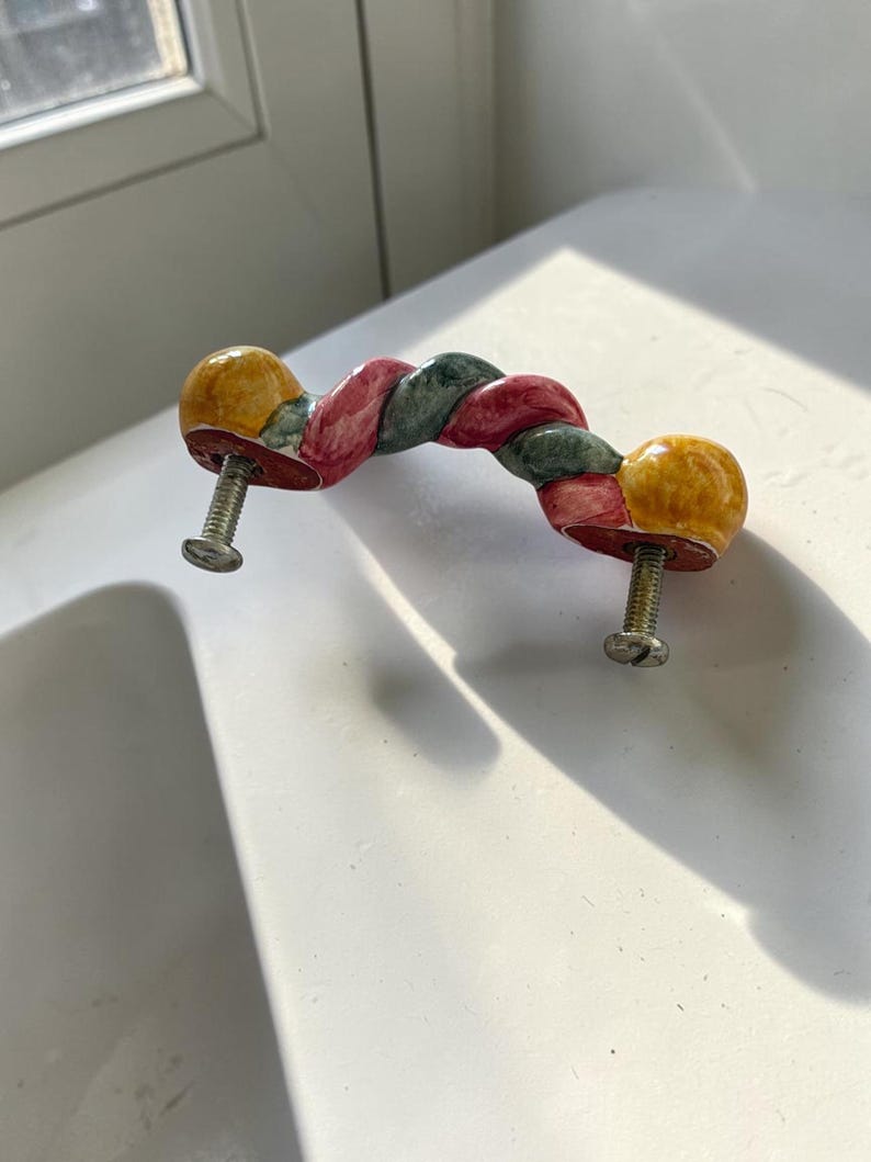 May include: A ceramic drawer pull featuring a twisted rope design with alternating segments of yellow, red, and dark green glaze. The handle has two metal mounting screws attached to the underside, extending outward for installation purposes.