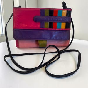 May include: A rectangular leather crossbody bag featuring a multi-colored design with panels of hot pink, purple, light pink, and a vertical striped section in red, orange, yellow, green, blue, and black. It has a thin black shoulder strap.