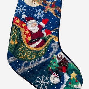 May include: A blue needlepoint Christmas stocking featuring a scene of Santa Claus in a green sleigh pulled by reindeer across a night sky with a full moon and snowflakes. The lower section depicts a house, a snowman, and a decorated pine tree.