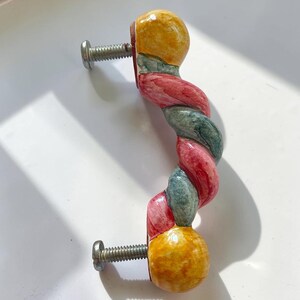 May include: A ceramic cabinet handle featuring a twisted, rope-like design with alternating segments of red and dark green, finished with solid yellow spherical ends. Two silver-toned metal mounting screws are attached to the back of the handle.