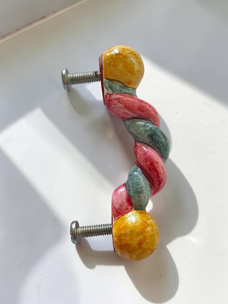 May include: A ceramic cabinet handle featuring a twisted, rope-like design with alternating segments of red and dark green, finished with solid yellow spherical ends. Two silver-toned metal mounting screws are attached to the back of the handle.