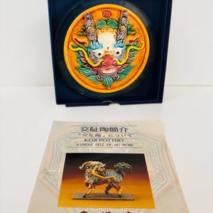 May include: A circular ceramic plaque featuring a colorful, relief-sculpted mythical creature face with horns, set in a dark blue box. Below is an informational card with the text "KOJI POTTERY A UNIQUE PIECE OF ART WORK" and an image of a creature.