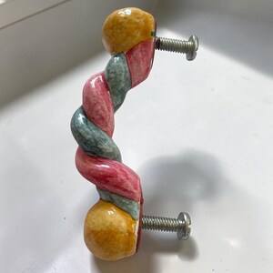 May include: A ceramic cabinet handle featuring a twisted, rope-like design with alternating segments of muted pink, sage green, and mustard yellow. The handle has two silver-toned metal screws extending from the back for mounting purposes.