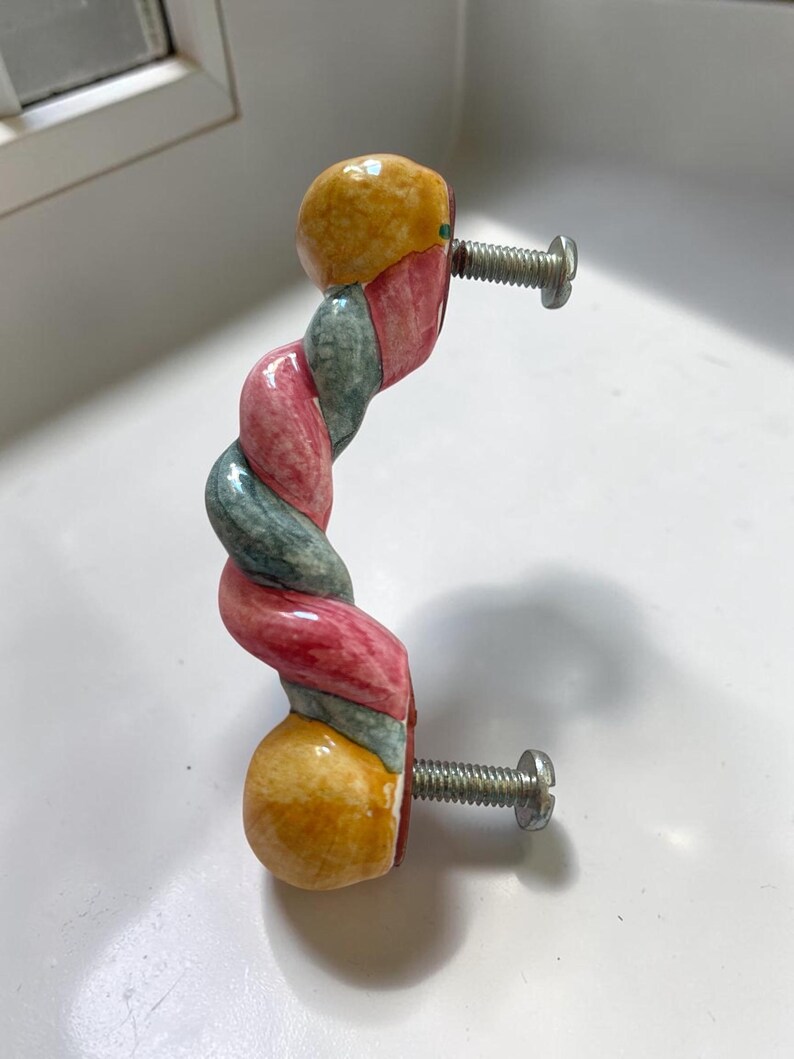May include: A ceramic cabinet handle featuring a twisted, rope-like design with alternating segments of muted pink, sage green, and mustard yellow. The handle has two silver-toned metal screws extending from the back for mounting purposes.