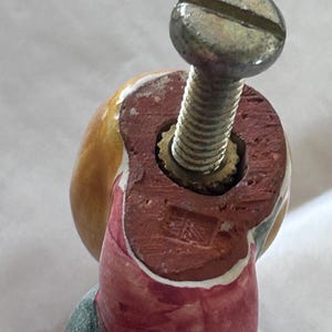 May include: A decorative ceramic knob featuring a rounded yellow top and a red base with a metallic screw protruding from the center. The base displays a small, square, stamped mark with the text "UK" visible on the flat, unglazed red surface.