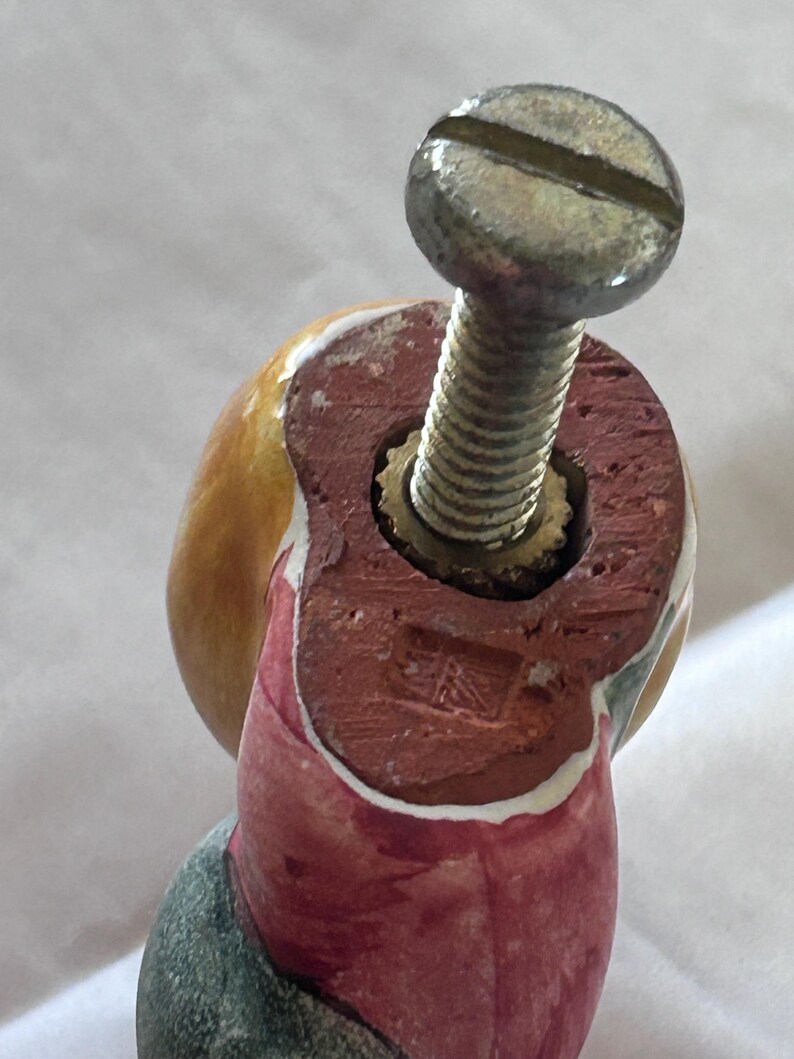 May include: A decorative ceramic knob featuring a rounded yellow top and a red base with a metallic screw protruding from the center. The base displays a small, square, stamped mark with the text "UK" visible on the flat, unglazed red surface.