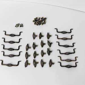 May include: A collection of antique brass-toned cabinet hardware including five long drawer pulls, twelve smaller knob-style pulls, a cluster of small circular metal fasteners, and several small screws arranged on a plain white surface.