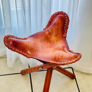 May include: A brown leather folding stool with a braided edge and wooden legs.