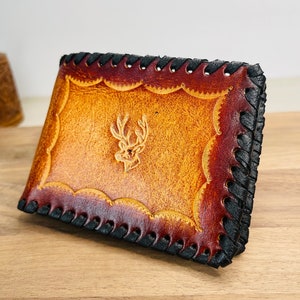 May include: Hand-stitched leather wallet with a deer head emblem. The wallet features a scalloped edge design and a gradient of brown and tan hues. The edges are stitched with black thread.