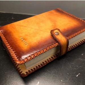 May include: A brown leather journal cover with a stitched edge and a strap closure. The cover has the name "DANIEL" embossed on the front.