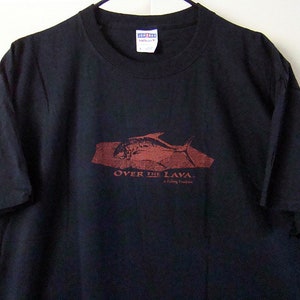 Może przedstawiać: Czarny T-shirt z czerwonym nadrukiem ryby pływającej nad skałą z tekstem "Over the Lava. A fishy thriller."