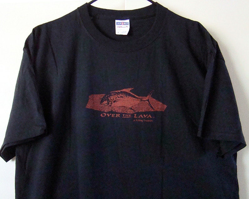 Może przedstawiać: Czarny T-shirt z czerwonym nadrukiem ryby pływającej nad skałą z tekstem "Over the Lava. A fishy thriller."