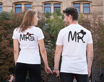 May include: A couple wearing white t-shirts with the words "MR." and "MRS." printed on the back. The shirts feature a black Mickey Mouse head with red ears.