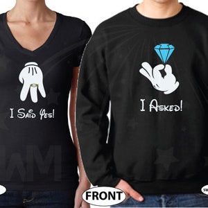May include: A black couple's matching t-shirt set. The woman's shirt says "I Said Yes!" with a cartoon hand wearing a ring. The man's shirt says "I Asked!" with a cartoon hand holding a blue diamond ring.