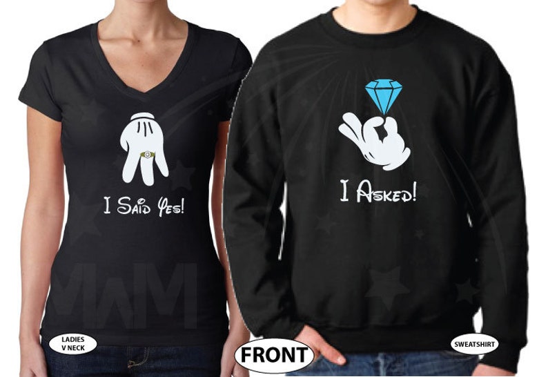 May include: A black couple's matching t-shirt set. The woman's shirt says "I Said Yes!" with a cartoon hand wearing a ring. The man's shirt says "I Asked!" with a cartoon hand holding a blue diamond ring.