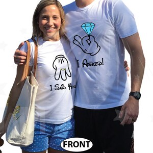 May include: A couple wearing white t-shirts with a Mickey Mouse hand graphic and the text "I Asked!" and "I Said Yes!"  The couple is standing in front of a white background.