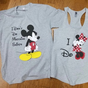 May include: A pair of gray t-shirts featuring Mickey and Minnie Mouse. The shirt with Mickey Mouse says "I Don't Do Matching Shirts". The shirt with Minnie Mouse says "I Do".