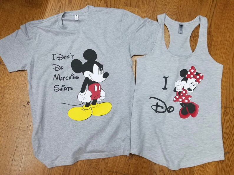 May include: A pair of gray t-shirts featuring Mickey and Minnie Mouse. The shirt with Mickey Mouse says "I Don't Do Matching Shirts". The shirt with Minnie Mouse says "I Do".