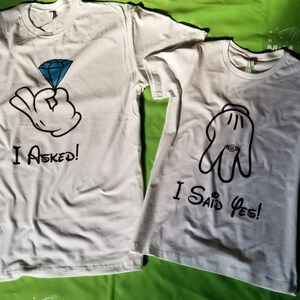 May include: Two white t-shirts with a cartoon hand design. The first shirt says "I Asked!" and the second shirt says "I Said Yes!"