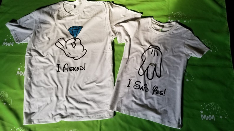 May include: Two white t-shirts with a cartoon hand design. The first shirt says "I Asked!" and the second shirt says "I Said Yes!"
