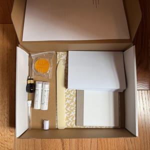 May include: A craft kit in a cardboard box containing a bone folder, two glue sticks, a small awl with a black handle, a clear plastic tube, a small orange circular tool, and a stack of white cardstock paper with a patterned yellow and white sheet.