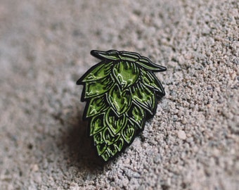 May include: A green enamel pin shaped like a hop cone. The pin has a black outline and is detailed with individual hop leaves.