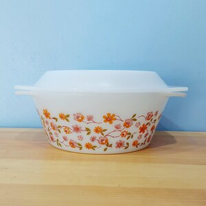 May include: White ceramic casserole dish with a floral pattern in shades of orange, pink, and green. The dish has a lid and is sitting on a wooden surface.